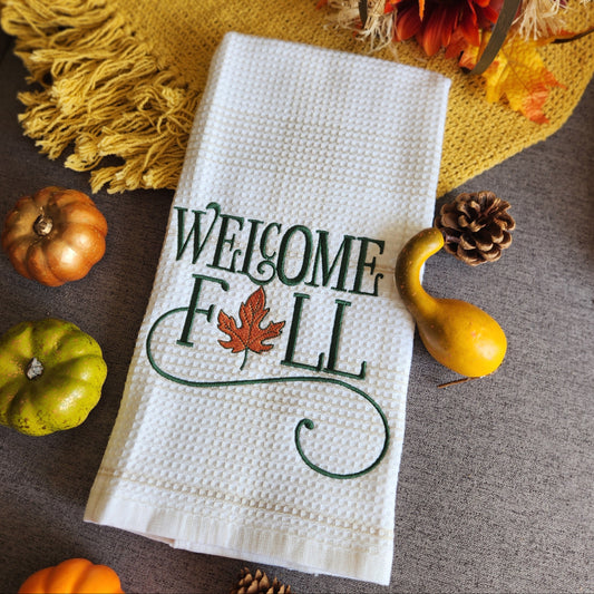 White towel with 'Welcome Fall' text and leaf design, surrounded by autumn decorations on a brown surface.